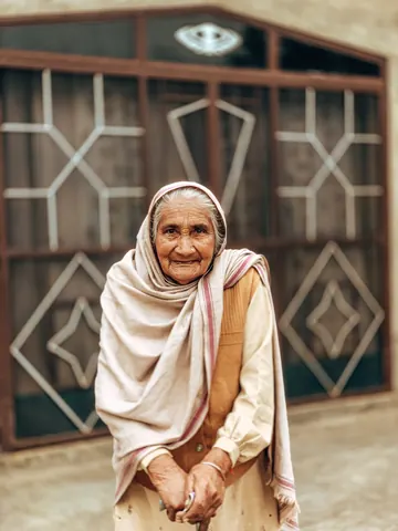 An elder stands with folded hands outside her home.